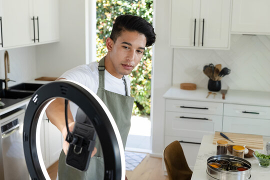 Man wearing green apron adjusting LED ring light and smartphone mount in modern white kitchen