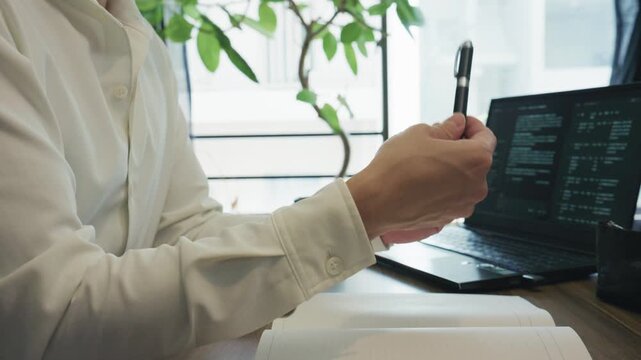 Side view of hands in white shirt turning a pen over open notebook at a desk