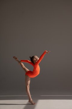 A little Caucasian girl gymnast in an orange leotard demonstrates her skills on a white background. 
