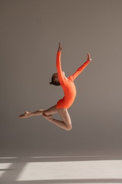 A little Caucasian girl gymnast in an orange leotard demonstrates her skills on a white background. 