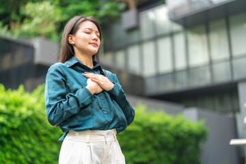 Fototapeta premium Mental health, happy young asian businesswoman taking a deep breath of fresh air with hand on chest standing outdoor, Relief, relaxing, corporate wellness, mindfulness and success concept
