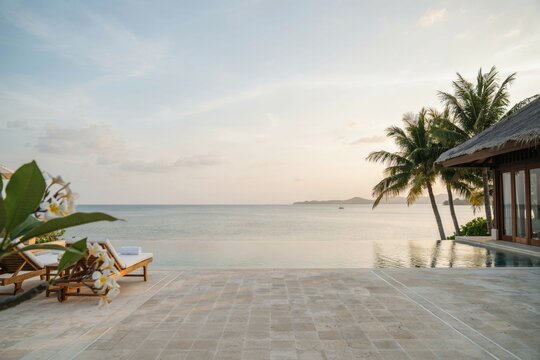 Infinity pool overlooking the calm ocean at sunset with palm trees, wooden lounge chairs, and white flowers on a tropical island resort deck