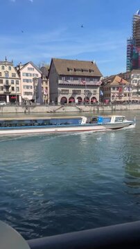 Perspective from a cafe terrace table with coffee, showing a passenger boat sailing on the Limmat River in Zurich, Switzerland, against the backdrop of historic architecture and Grossmunster cathedral