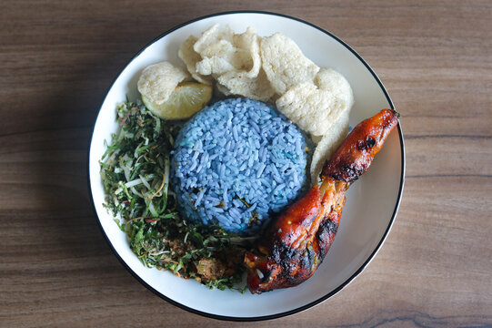 Top-down view of a plate of Malaysian Nasi Kerabu featuring blue pea flower rice served with grilled chicken, raw vegetable and herb salad (ulam), and fish crackers.