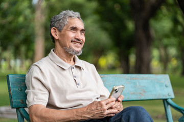 Fototapeta premium portrait elderly pensioner man spend leisure time relaxing in urban park,sitting on bench,holding mobile phone,smiling and looking in nature