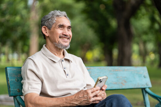 portrait elderly pensioner man spend leisure time relaxing in urban park,sitting on bench,holding mobile phone,smiling and looking in nature