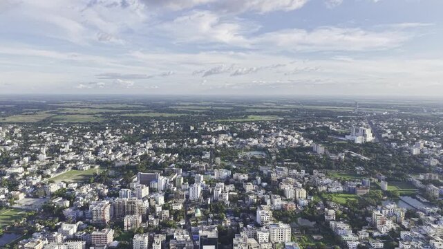 Drone footage overlooking Kushtia, Bangladesh, captures a broad urban landscape with residential buildings, smaller structures, dense green vegetation, and clouds across the blue sky.