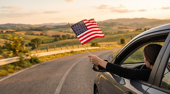 Woman waving American flag from car window on scenic rural road during sunset.