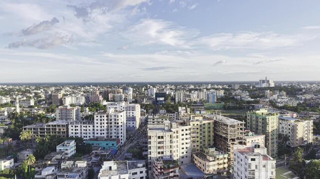 Drone camera moves upward with city buildings in the foreground, revealing more of Kushtia's urban area, surrounding greenery, and cloudy blue sky.