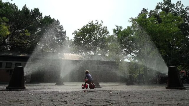 slow motion girl on tricycle rides through sprinklers in Brooklyn park