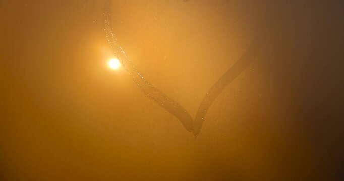 The bright yellow sun outside the window, covered with condensation, the setting sun shines through small liquid drops on the glass and the heart pattern