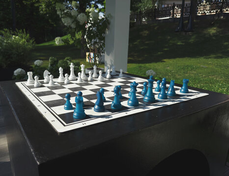 Large outdoor chess set with white blue pieces arranged on stone table, park garden background, strategy game concept for leisure, business competition