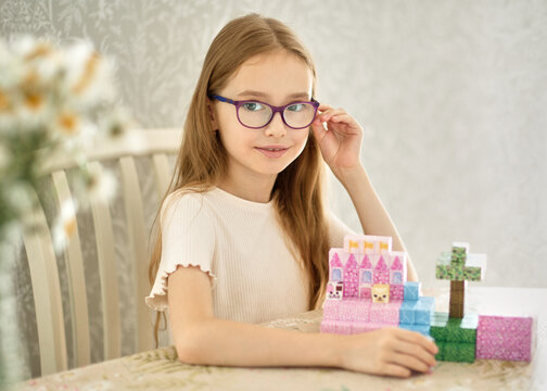 Smiling girl with glasses holds a colorful magnetic blocks castle she is building while playing at home, enjoying a creative childhood activity with construction toys in a bright cozy interior.