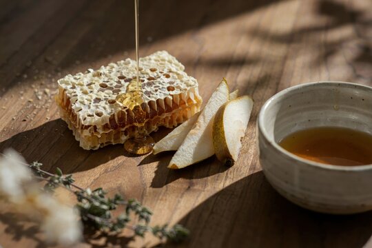 Golden honeycomb dripping with liquid honey next to fresh pear slices and a small bowl of honey on a rustic wooden cutting board in sunlight