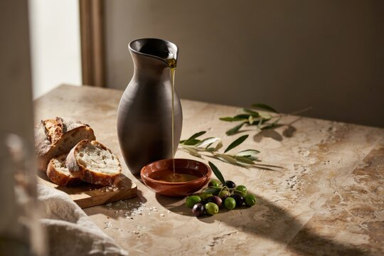 Golden olive oil pouring from a dark ceramic pitcher into a small bowl, with rustic sourdough bread slices and fresh green olives on a table