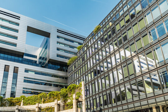 View of modern office buildings with glass facades and metal cladding, featuring a unique architectural cutout and green plants on a stone wall in London, England, United Kingdom.