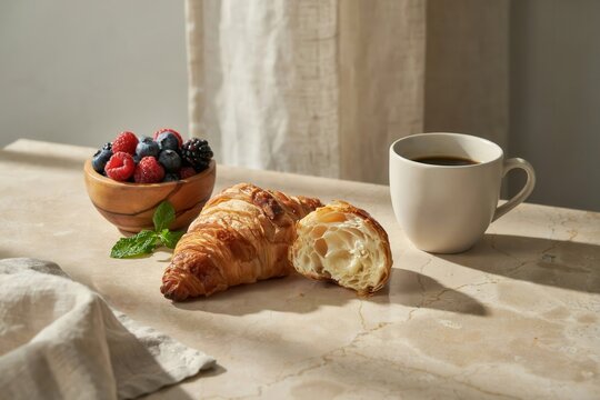 A buttery croissant sliced in half next to a wooden bowl filled with fresh raspberries, blueberries, and blackberries with a cup of black coffee