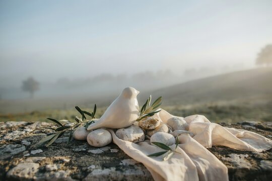 A ceramic white bird figurine resting on weathered stone with olive branches and soft fabric against a misty morning landscape background