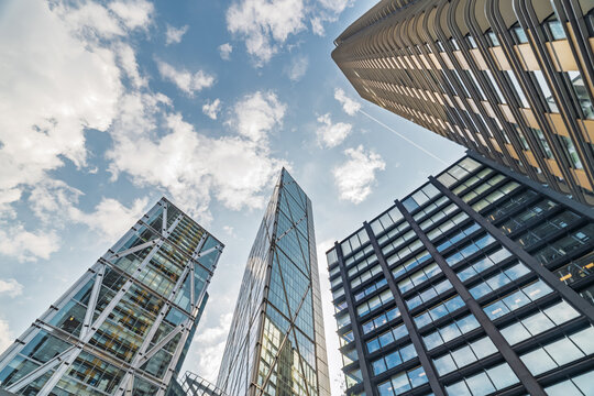 View of modern skyscrapers with glass facades and steel structures reaching towards a blue sky with scattered clouds in London, England, United Kingdom.