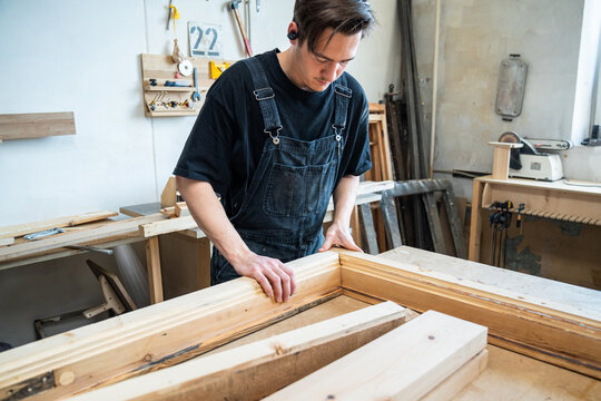 Craftsman connecting parts of refurbished antique wooden window frame at workbench in workshop, focused handicraftsman accurately putting together all details of reconstructed frame at workstation.