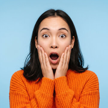 Shocked young woman wearing orange sweater looking amazed with hands on cheeks against blue