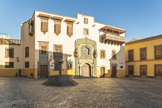 View of the historic Casa de Colon with its ornate stone portal and fountain in the cobblestone square of Las Palmas de Gran Canaria, Canary Islands, Spain.