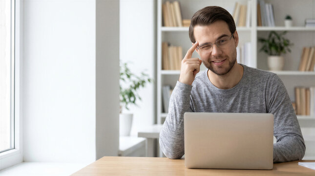 Portrait of a smiling young man sitting at a desk with a laptop, touching his glasses and winking at the camera in a home office