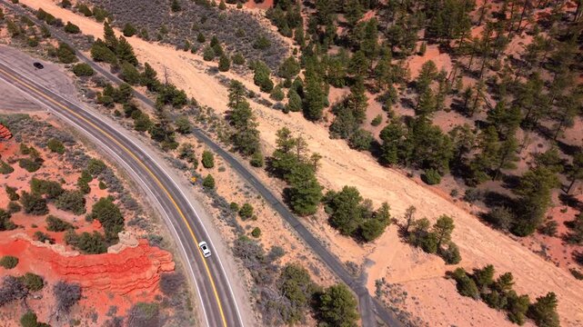 Aerial drone footage of Utah Scenic Byway 12 showcasing cars traveling along a curved desert highway. Red rock formations and green juniper trees dot the arid landscape under bright sunlight.