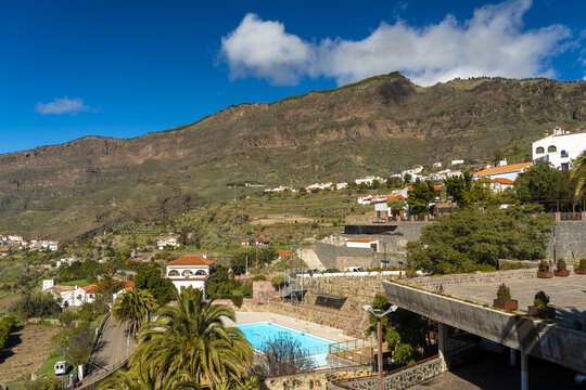 View of the village of Tejeda nestled in the mountainous landscape of the Canary Islands with a swimming pool and palm trees in the foreground, Tejeda, Canary Islands, Spain.