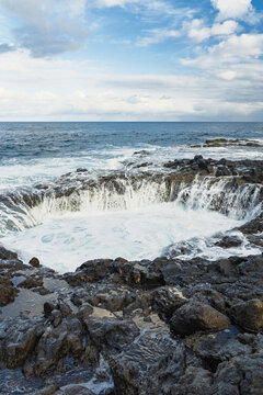 View of the natural blowhole at La Garita with crashing ocean waves, volcanic rock formations, and sea spray under a blue sky in La Garita, Canarias, Spain.