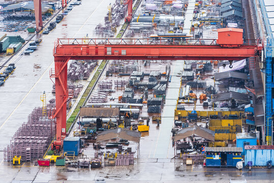 Busy shipbuilding yard with workers carrying out construction activities under enormous overhead cranes among large steel hull sections, pipes and industrial materials.