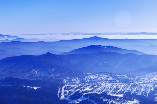 Panoramic winter view of Sheregesh ski resort in Russia see from snowy mountain Mystag slope. Snow-covered forest, village and hotels, distant peaks on horizon under clear blue sky, tonal perspective