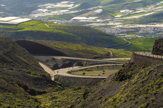 View of the winding road through the volcanic mountains of La Aldea de San Nicolas with green hills and greenhouses in the distance in Las Palmas de Gran Canaria, Canary Islands, Spain.