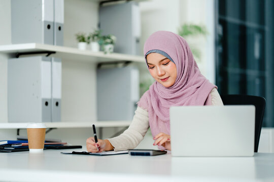 Women wearing hijab working on laptop and writing note in office