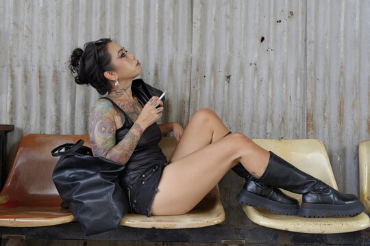 Asian tattoo female model in a black tank top and denim shorts sits holding a cigarette