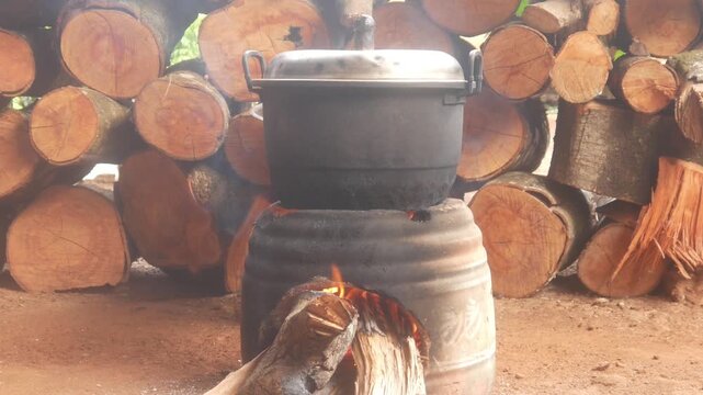 Traditional cooking using a large black pot on a wood burning stove, with a background of stacked logs in a rural kitchen.