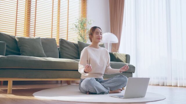 Happy young Asian woman practicing yoga and meditation at home sitting on floor in living room in lotus position with laptop