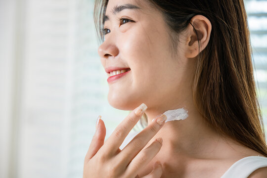 Asian beautiful young woman applying cosmetic cream on neck at home