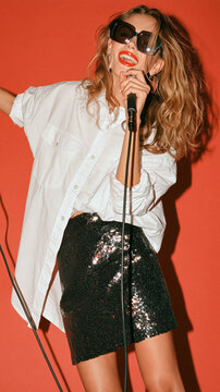 Rockstar on Stage: A confident woman with sunglasses and red lipstick sings passionately into a microphone, her curly hair and sequined skirt adding to her vibrant stage presence.