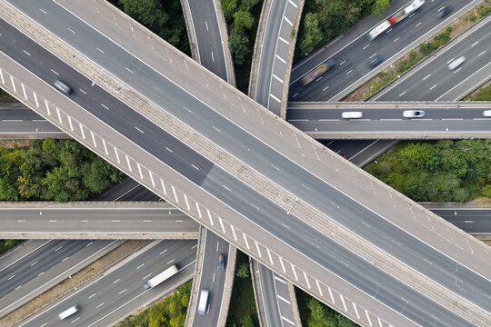 Aerial view of a complex highway interchange with multiple lanes of traffic and vehicles moving across concrete overpasses surrounded by dense green trees in the United Kingdom.
