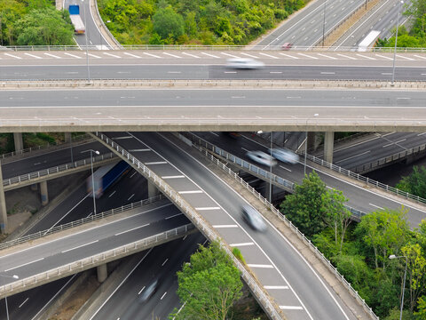 Aerial view of a complex highway interchange with multiple levels of roads, moving vehicles, and surrounding green trees in the United Kingdom.