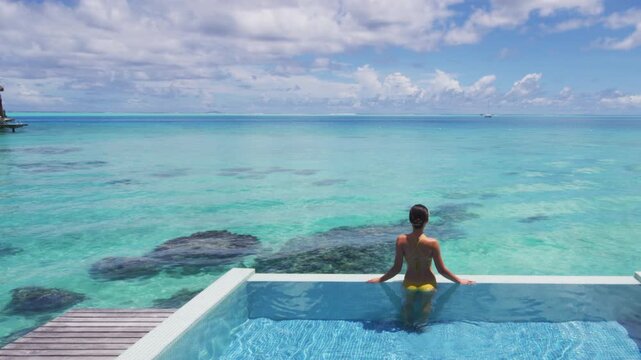 Summer sunbathing happy bikini woman swimming in infinity pool of luxury resort hotel relaxing in idyllic overwater ocean view suite. Suntan solar skin care. Raw unedited video of real woman model.