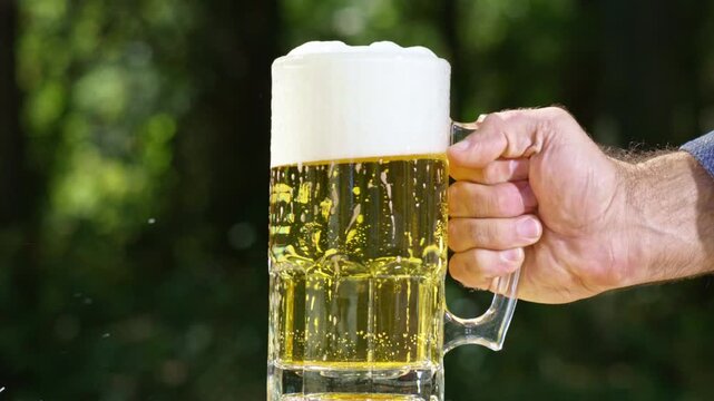 Hand holding beer mug outdoors, Hand with beer glass on green nature background. Beer and summer drink. Beer splash in slow motion.
