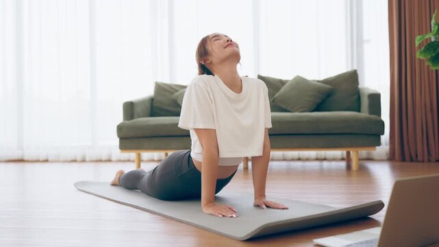 Young Asian woman stretching yoga workout on exercise mat while online training class with computer laptop in living room