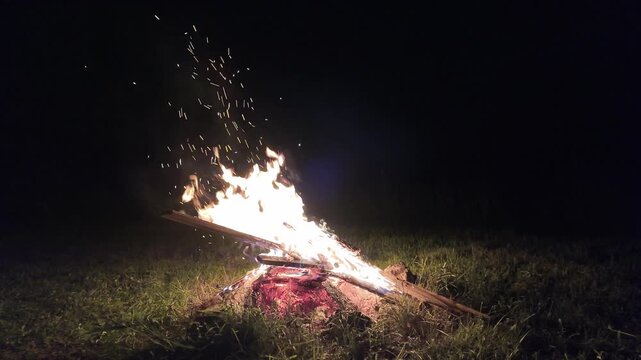 Cozy bonfire blazing on a grassy meadow at night, casting warm firelight and glowing sparks into the darkness. Perfect for camping, hiking, wilderness and outdoor adventure themes.