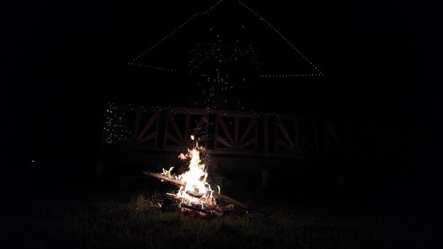 Cozy bonfire blazing on a grassy meadow at night, casting warm firelight and glowing sparks into the dark.