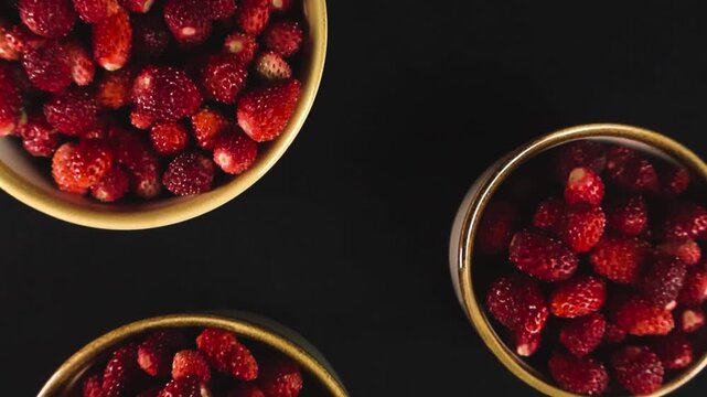 Two ceramic bowls full of freshly picked ripe red wild strawberries slowly rotating on a black background, representing a healthy and organic vegetarian summer dessert from the forest