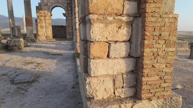 Ground-level cinematic view of the Roman Basilica at the UNESCO-listed Volubilis archaeological site, highlighting monumental arches and preserved Roman judicial architecture in Morocco.