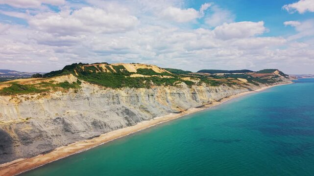 Sweeping aerial drone footage travelling beside the dramatic eroded cliffs, narrow beach and turquoise sea near Charmouth on Dorset&rsquo;s Jurassic Coast, England. The coastal view highlights landslip