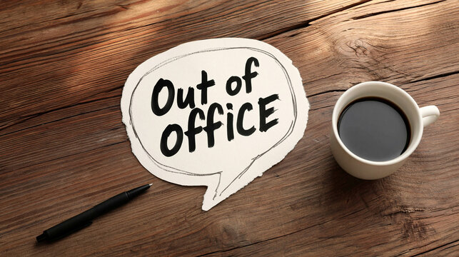 Out of Office Notification: A speech bubble with the words Out of Office written on it rests beside a pen and a cup of coffee on a rustic wooden table, symbolizing a break from work.
